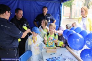 trzej policjanci w namiocie promocyjnym z grupką dzieci, na stoliku policyjne gadżety oraz przymocowane nadmuchane balony