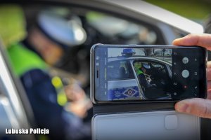 Ekran telefonu komórkowego, na którym widać policjanta siedzącego w radiowozie.