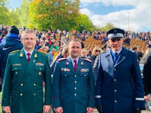 Na pierwszym planie trzej mundurowani policjanci pozują do zdjęcia.Od lewej strony stoją dwaj oficerowie Policji litewskiej a z prawej generał z Polski. W tle widać ludzi - jedni siedzą, inni stoją.