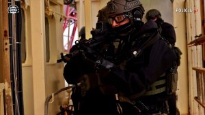 Policyjni uzbrojeni kontrterroryści w specjalistycznym umundurowaniu, w kaskach i okularach znajdują się w wąskim jasnym korytarzu, trzymając broń w gotowości do ataku.