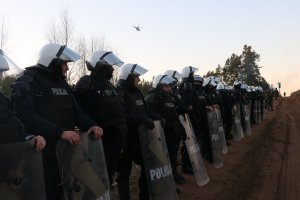 umundurowani policjanci z tarczami i w białych kaskach stoją wzdłuż drogi na granicy. Nad nimi lata policyjny śmigłowiec