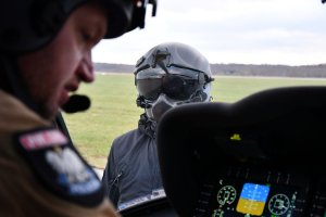 widok na kokpit w policyjnym śmigłowcu, na część twarzy pilota i stojącego poza śmigłowcem mężczyzny w skafandrze i kasku