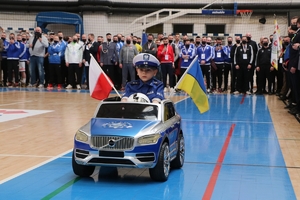 mały chłopiec w policyjnym mundurze jedzie w dziecięcym samochodzie przez halę sportową, w tle uczestnicy turnieju