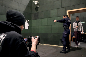 policjantka na strzelnicy celuje z broni, z przodu i z tyłu widać dwóch mężczyzn, jeden z aparatem fotografuje policjantkę, drugi nagrywa materiał kamerą