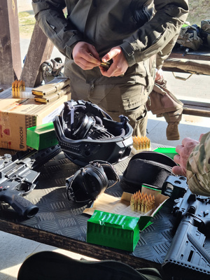 Metalowy stół, na którym znajdują się m.in.: broń, amunicja, nauszniki, załadowane magazynki. W drugim planie zdjęcia umundurowany policjant ładujący magazynek amunicją.