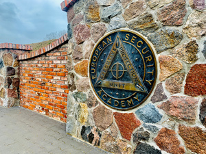 Kamienny mur z wmurowaną okrągłą mosiężną tablicą, na niej napisy European Security Academy i Executive Protecion Unit.