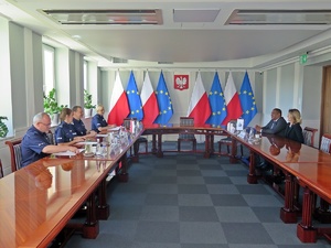 czworo polskich oficerów siedzi za stołem, naprzeciwko siedzi mężczyzna w garniturze i kobieta. Z tyłu widoczne są ustawione  w stojakach flagi