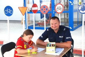 uśmiechnięty policjant siedzi z dziewczynką przy stoliku, dziewczynka rysuje