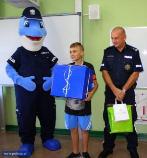 dwaj policjanci, jeden przebrany za maskotę policyjną, drugi stoi z prezentem a między nimi stoi chłopiec
