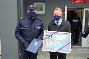 Komendant Wojewódzki Policji w Bydgoszczy trzyma teczkę z logiem Bydgoszczy i stoi obok mężczyzny z urzędu miasta, który trzyma w rękach obraz