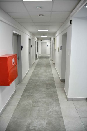 Jasny korytarz, ściany białe. Na podłodze płytki jasnoszare po bokach i ciemnoszare ułożone  pasem przez środek korytarza. Po prawej, po lewej stronie i na wprost na końcu korytarza zamknięte drzwi do pomieszczeń. Obok nich granatowo-białe tabliczki. Po lewej stronie hydrant wewnętrzny w czerwonej, metalowej szafie. Ściany białe. sufit jasny, kasetonowy z wpuszczanymi panelami Led.