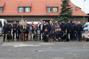 Zdjęcie grupowe przewodników psów ze swoimi czworonogami wraz z uczestnikami szkolenia - na zdjęciu kilkadziesiąt osób, w tle budynki.