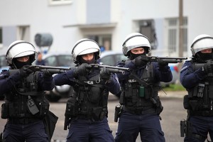 Czterech policjantów w czarnych strojach do działań pododziałów zwartych i białych kaskach stoją mierzą przed siebie z długiej broni.