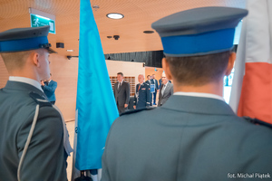 Zdjęcie przedstawia stojących tyłem policjantów z flagami Interpolu i Polską, w tle widać Sekretarza Interpolu,Komendanta Głównego Policji i Wiceprezydenta Interpolu