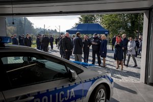 zdjęcie kolorowe: na pierwszym planie oznakowany radiowóz policyjny, w tle policjanci oraz goście biorący udział w uroczystości