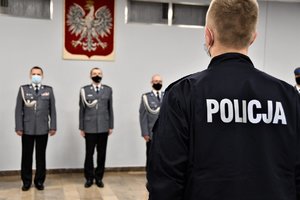 Na pierwszym planie plecy nowo przyjętego policjanta - widoczny napis Policja, w tle stojący frontem do fotografa przedstawiciele kierownictwa śląskiej Policji.