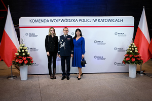 Zdjęcie przedstawia policjanta oraz dwie kobiety stojących na tle napisu Komenda Wojewódzka Policji w Katowicach oraz dwie flagi Polski